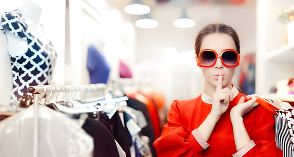 Seorang individu mengenakan pakaian merah mencolok dan kacamata besar berdiri di toko pakaian yang terang, melakukan gestur "diam" dengan jari di bibir, dikelilingi rak baju dan manekin.